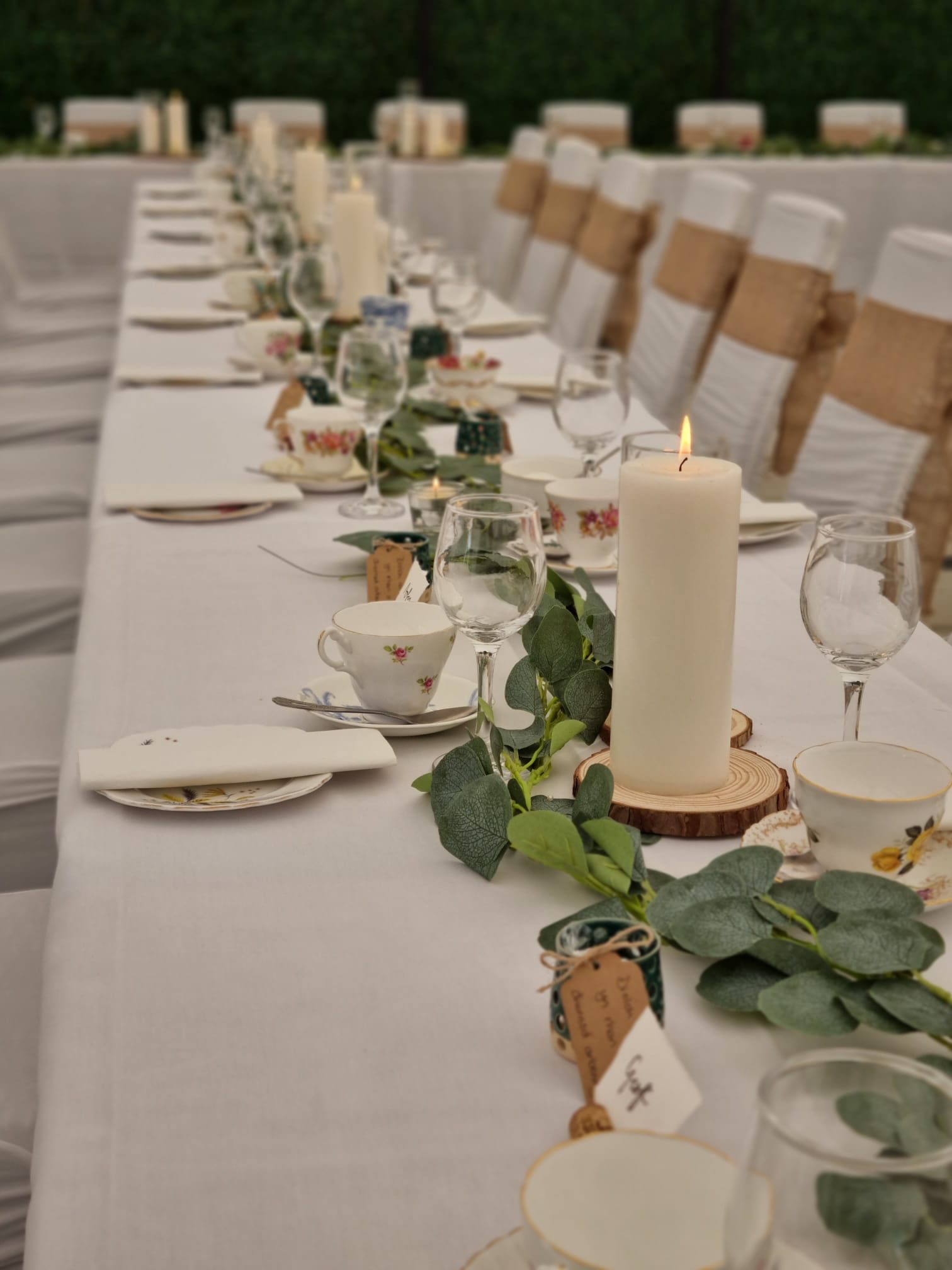 A long wedding breakfast table laid with white linen, candles, vintage china and a eucalyptus runner