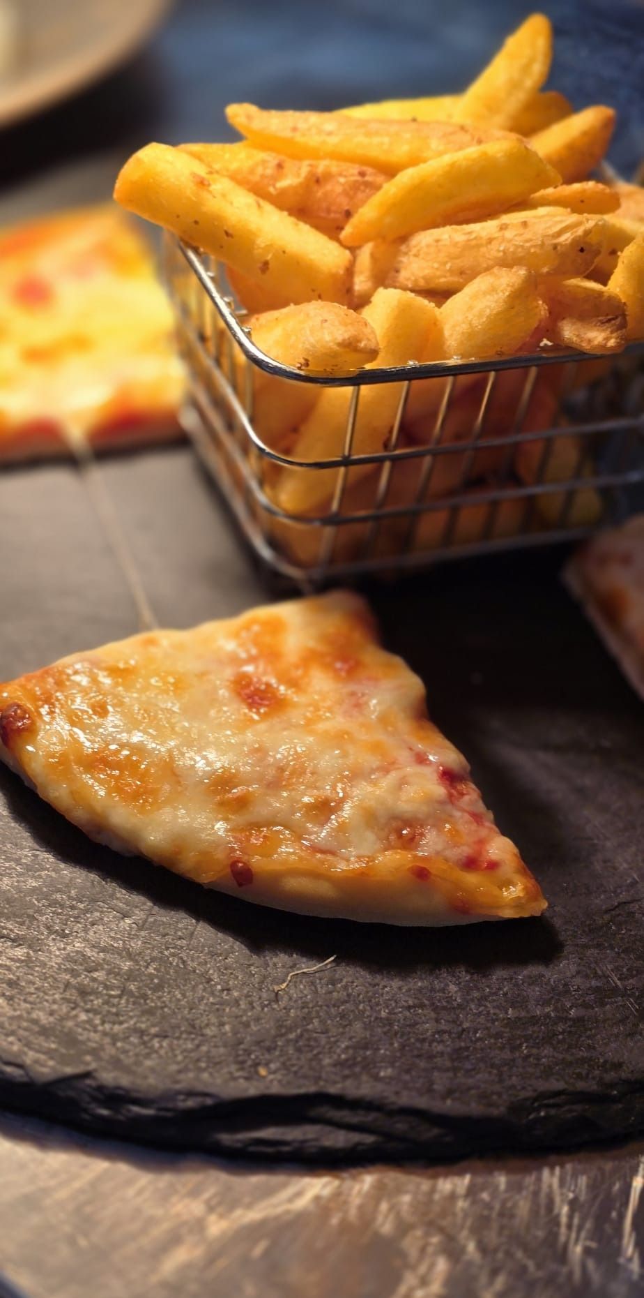 A cheese and tomato pizza slice on slate with a wire basket of chips