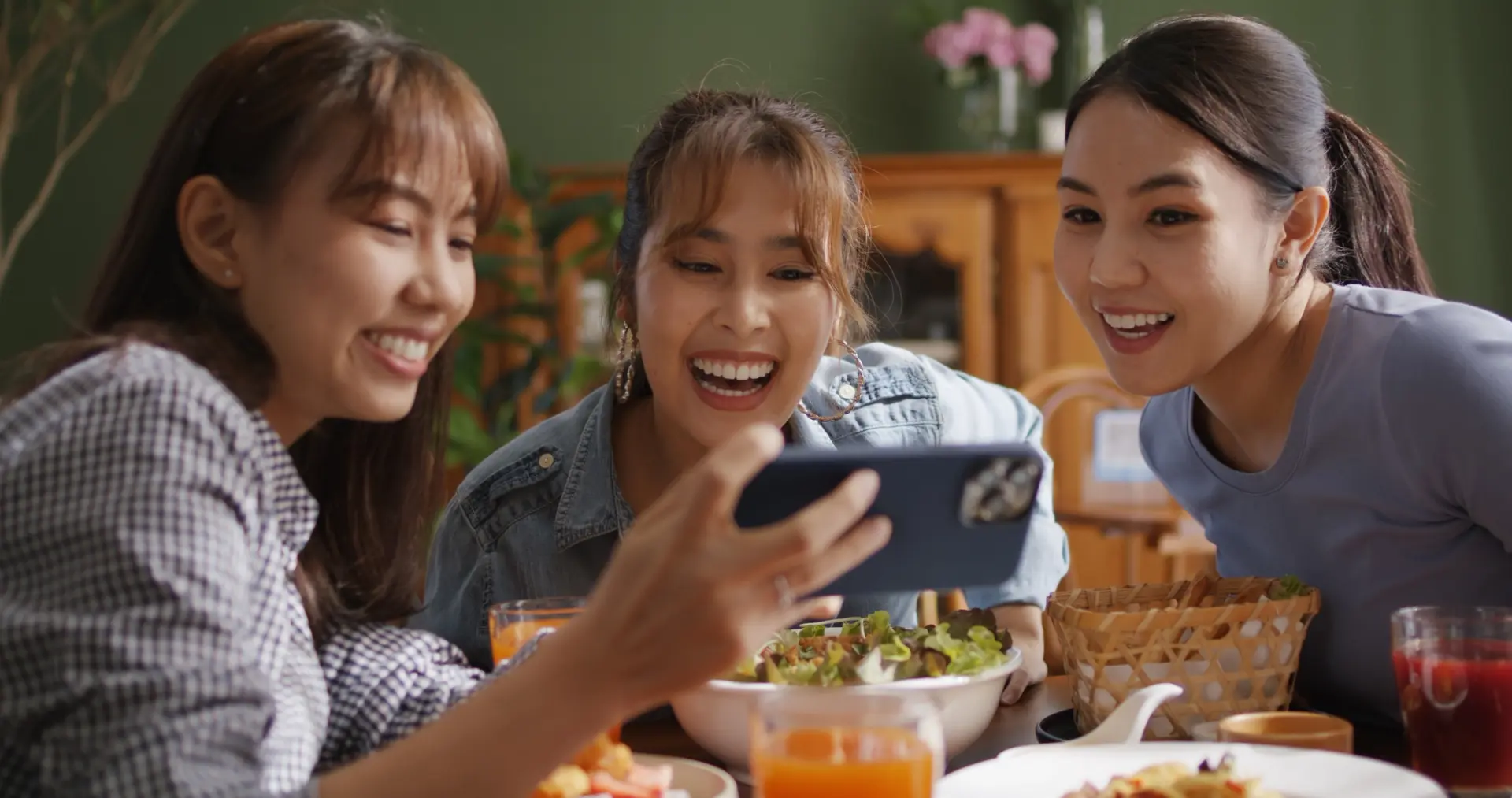 Tres amigas riendo ante la pantalla de un móvil en una mesa con comida y bebidas