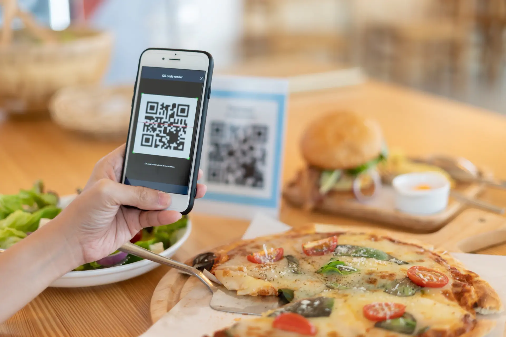 يد تمسح رمز QR على الطاولة بجانب بيتزا وبرغر وسلطة