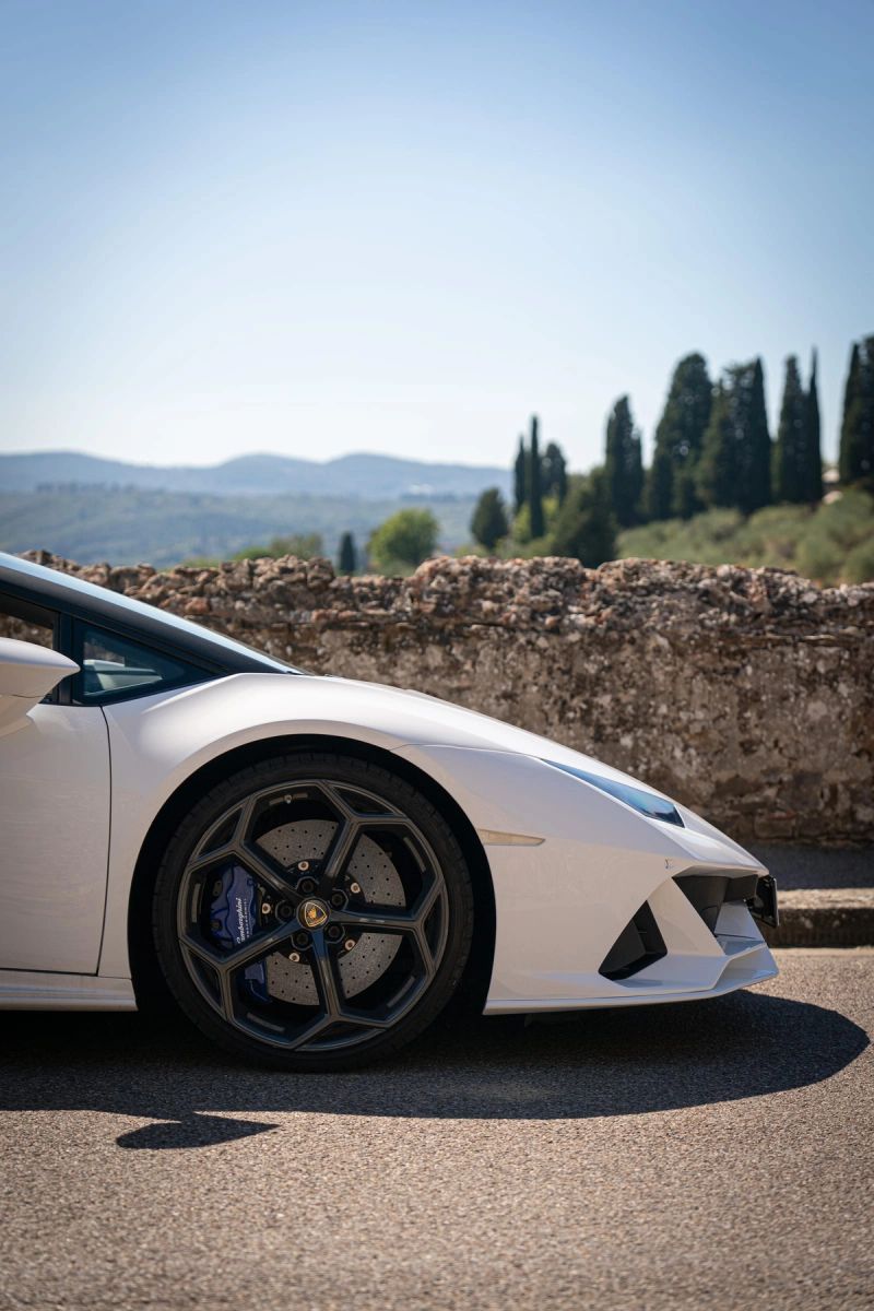 Una Lamborghini bianca ferma su una strada di campagna, fra cipressi e un muro a secco