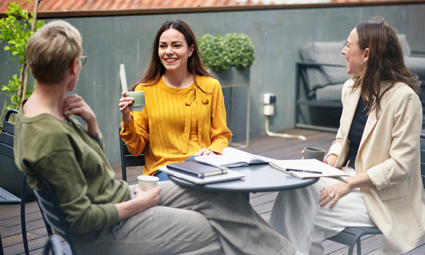 The main image for this comundo blog post about working a four-day workweek shows three women sitting at a small round table. They are relaxed and smiling and have cups of tea or coffee in their hands. There are closed laptops and notebooks on the table suggesting a relaxed work scene