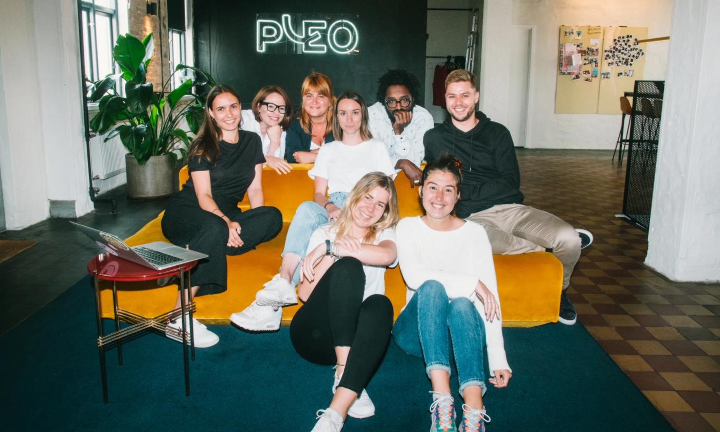 A group of smiling Pleo employees sitting on a yellow sofa with a neon Pleo sign on the wall behind them