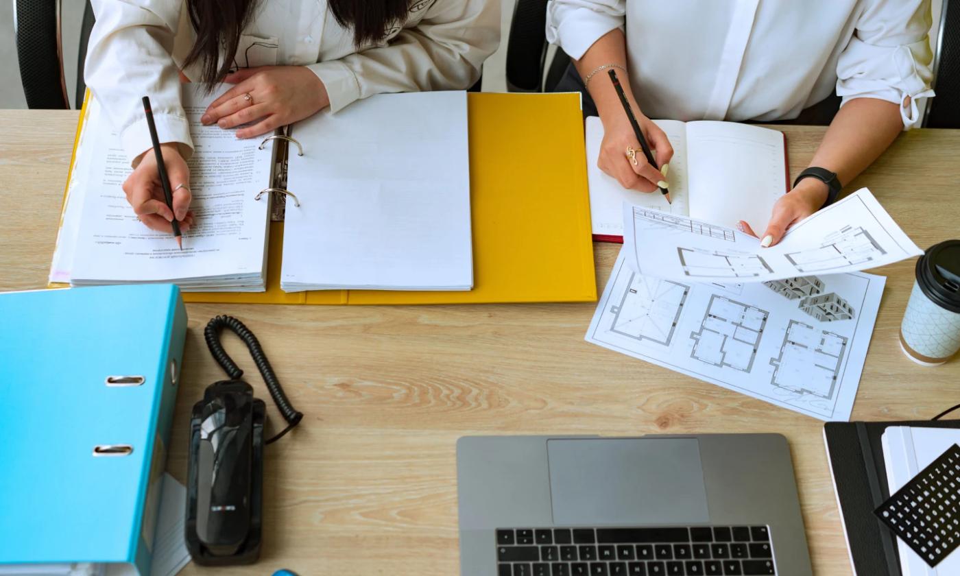 The main image of this comundo blog post about how the EPC is becoming a financial metric shows two people working at a desk. We see it from above, and can only see their hands. On the table is a laptop, some paper and a large file