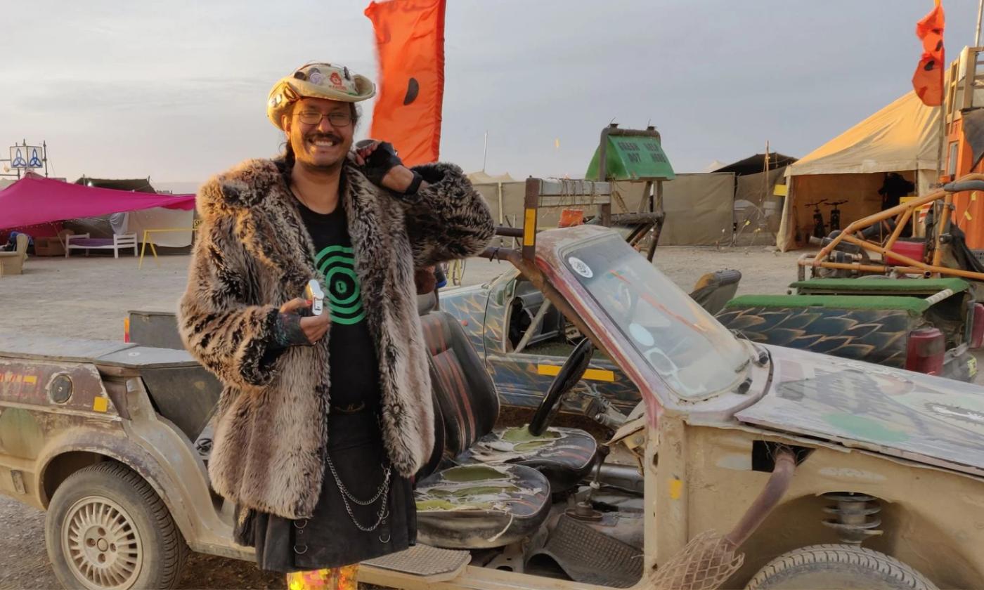 This is a photo of Brad standing by a buggy at Burning Man Festival. There are tents and flags in the background, and he looks very happy