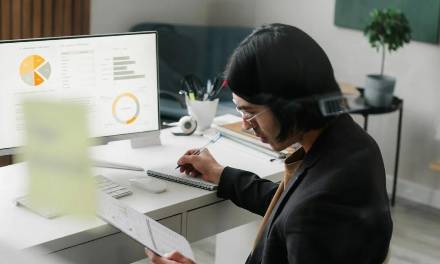 This comundo blog post about CSRD has a header image of a man sitting at an office desk, with a desktop monitor, keyboard and mouse on it. He's holding some paper, looking at it, and taking notes with his other hand. On the computer screen we can see various charts.
