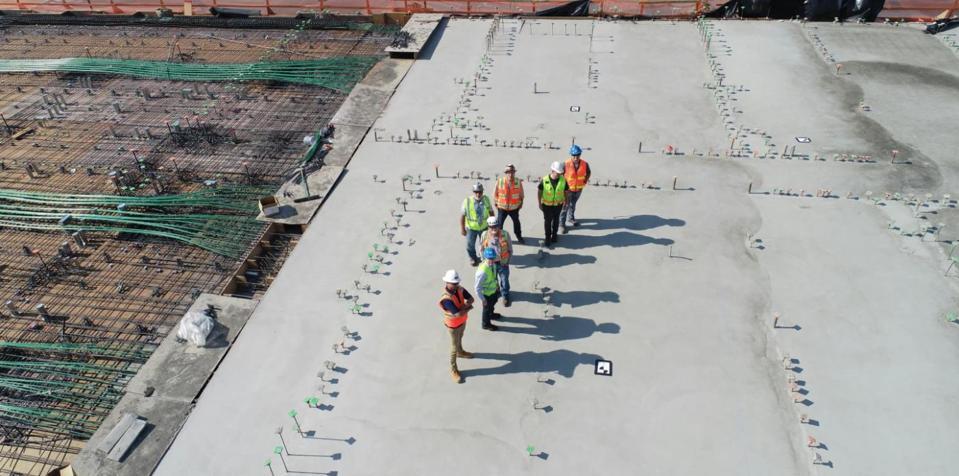 A low aerial shot of group of construction workers in high-vis jackets and hard hats are standing on top of what looks like the base of a building. They are standing on concrete but to the left there is no concrete yet, and we can see cables and iron poles. 