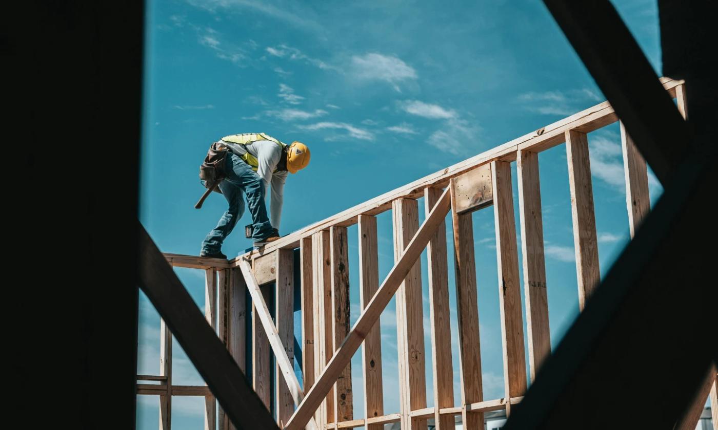 This comundo blog post about DGNB certification has a main image showing a construction worker standing on a wooden frame of a building. He's bending over and appears to be working on the frame. The sky is blue in the background.