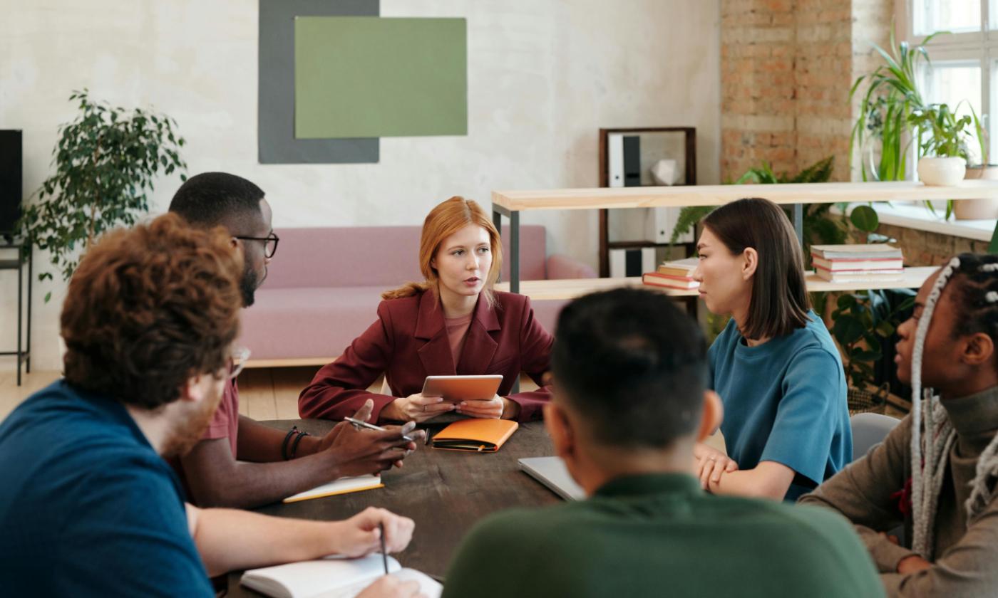In this photo we see a diverse group of young people sitting at a table during a meeting. A woman seems to be leading the meeting, and the others have notebooks on the table and are listening intently. 