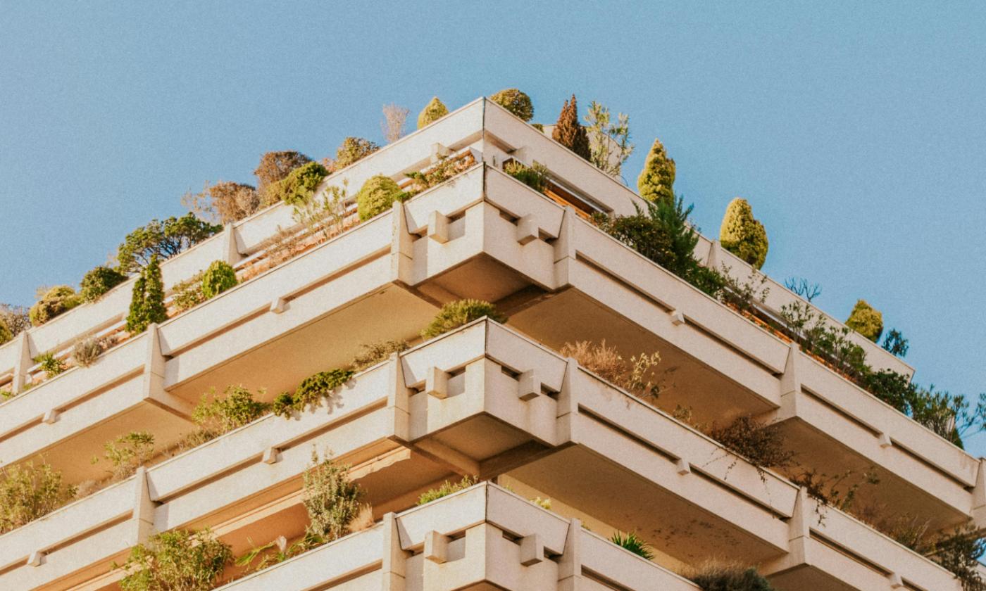 The main image for this comundo blog post about the NABERS rating shows a corner of a building taken from a lower perspective. This makes it look a little triangular. The sky behind is clear and blue and the building a sandy colour, with plants hanging off balconies that seem to wrap around the building.