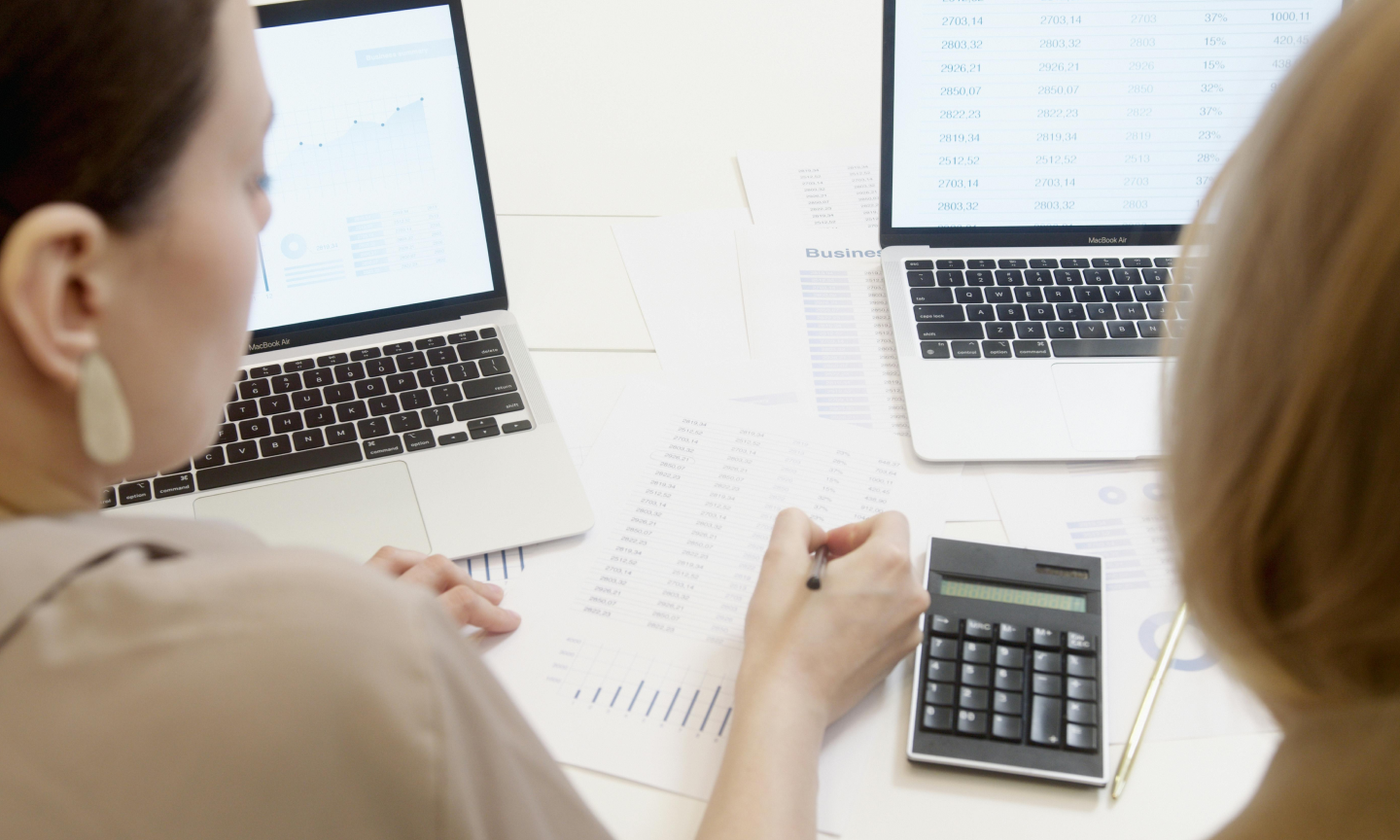 This comundo blog post about the cost of not automating energy reporting shows two women pouring over invoices. Our view is behind them, we have a partial view of the backs of their heads, and we can see a calculator, some printed invoices and two laptops open on the table.