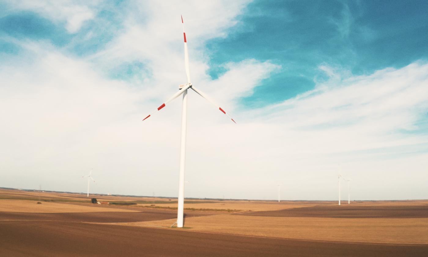 In this comundo blog post about Deepki and comundo's partnership, the main image is a photo of a wind turbine. The turbine is in an expanse of flat ploughed fields. We can see some more turbines in the distance, and the sky is blue with light, white clouds.