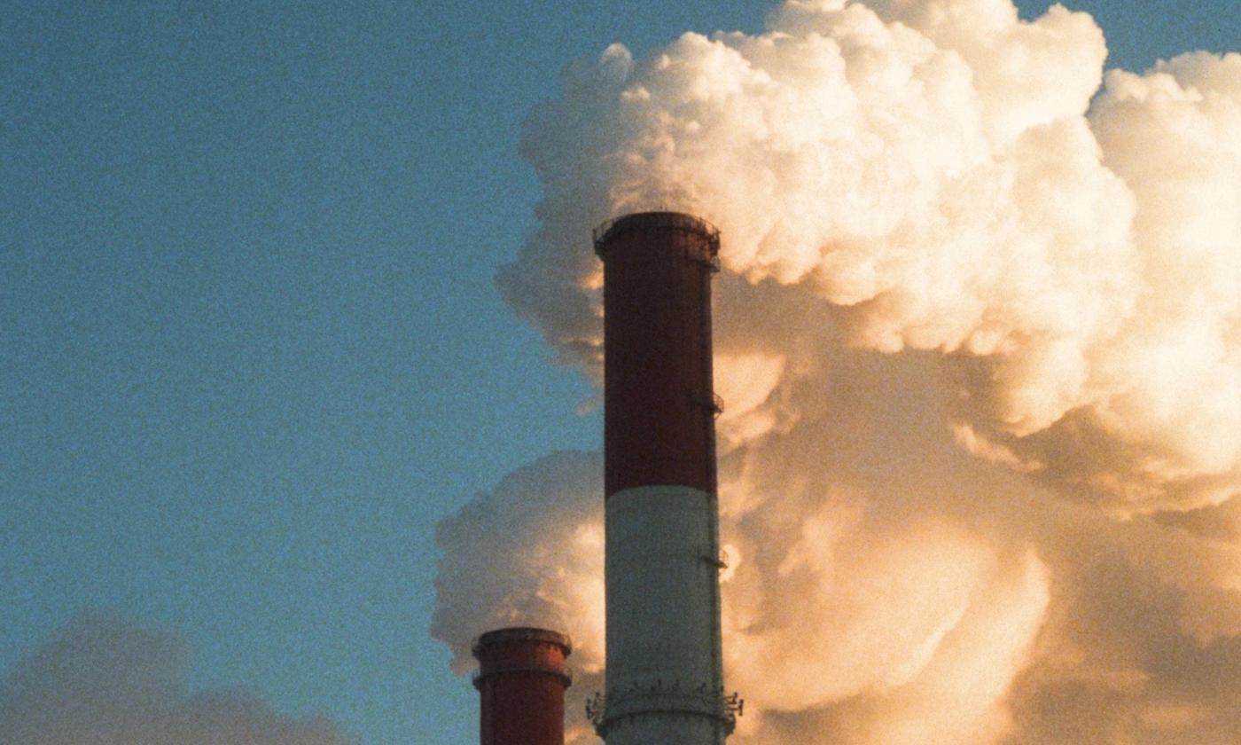 The main image for this comundo blog post about carbon tax shows the tops of two smoke stacks. They are both billowing out steam into a dark but blue sky