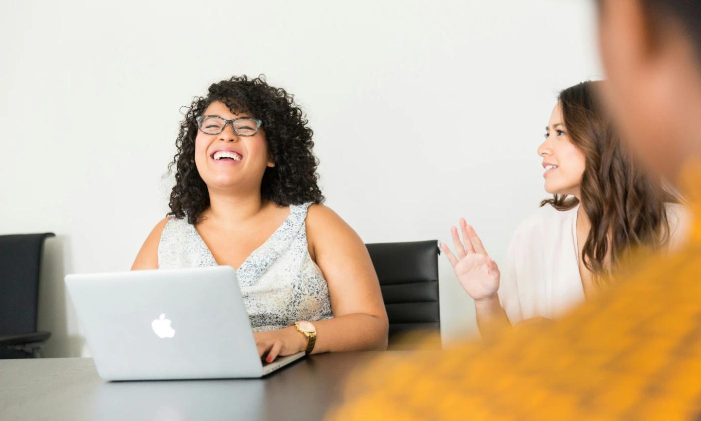 We're looking at a woman of African American appearance who is sitting at a table with her hands on an open laptop, laughing with her eyes close and head tilted back. We're looking over the shoulder of someone we can't really make out, and can see another woman who looks as though she's saying something that's making the first woman laugh