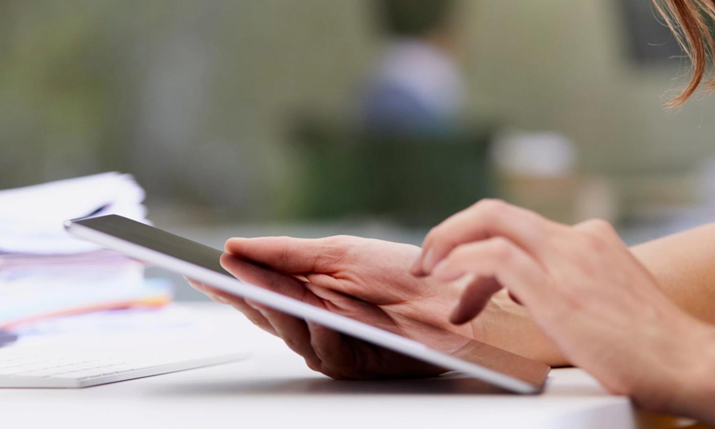 A close-up photo showing a woman's hands using a tablet with a pile of paper on the table she's sitting at