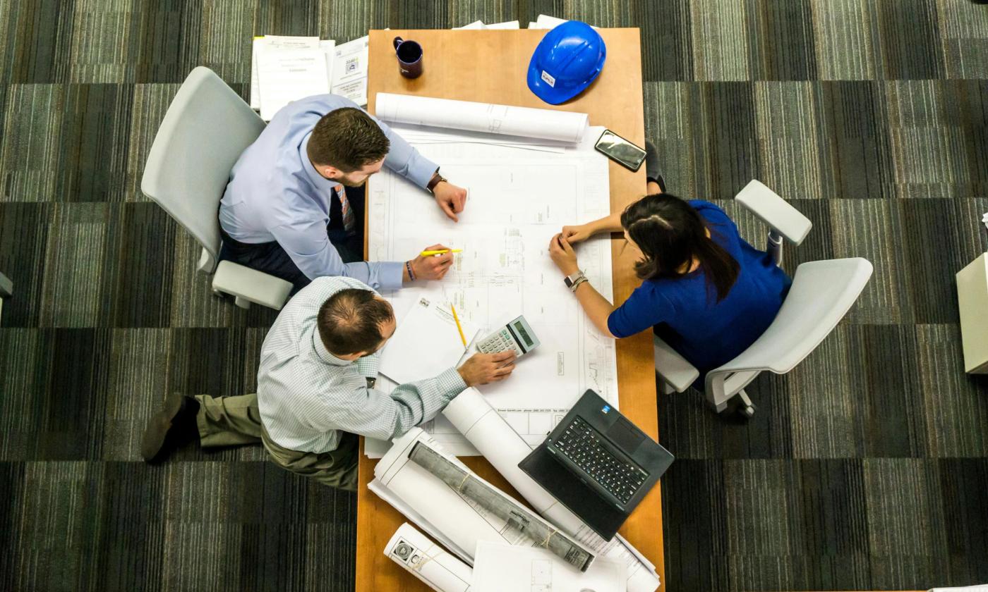 This comundo blog post about green financing in real estate is of three people sat around a table that is in a workplace. They have blueprints, laptops and calculators out. It is a bird's eye view so we can't see faces.