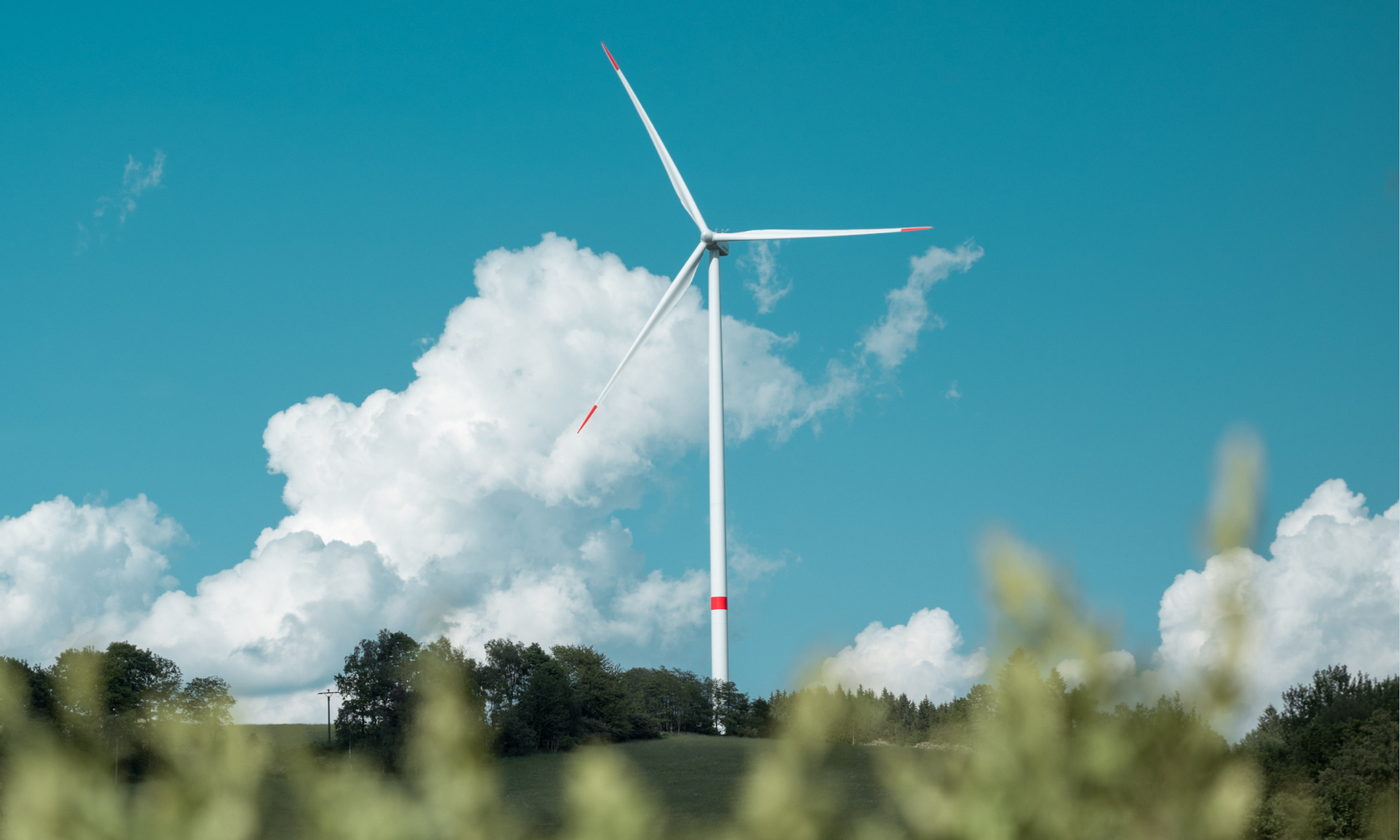 A photo of a slender wind turbine with blue sky and white clouds behind it, and with greenery at the forefront