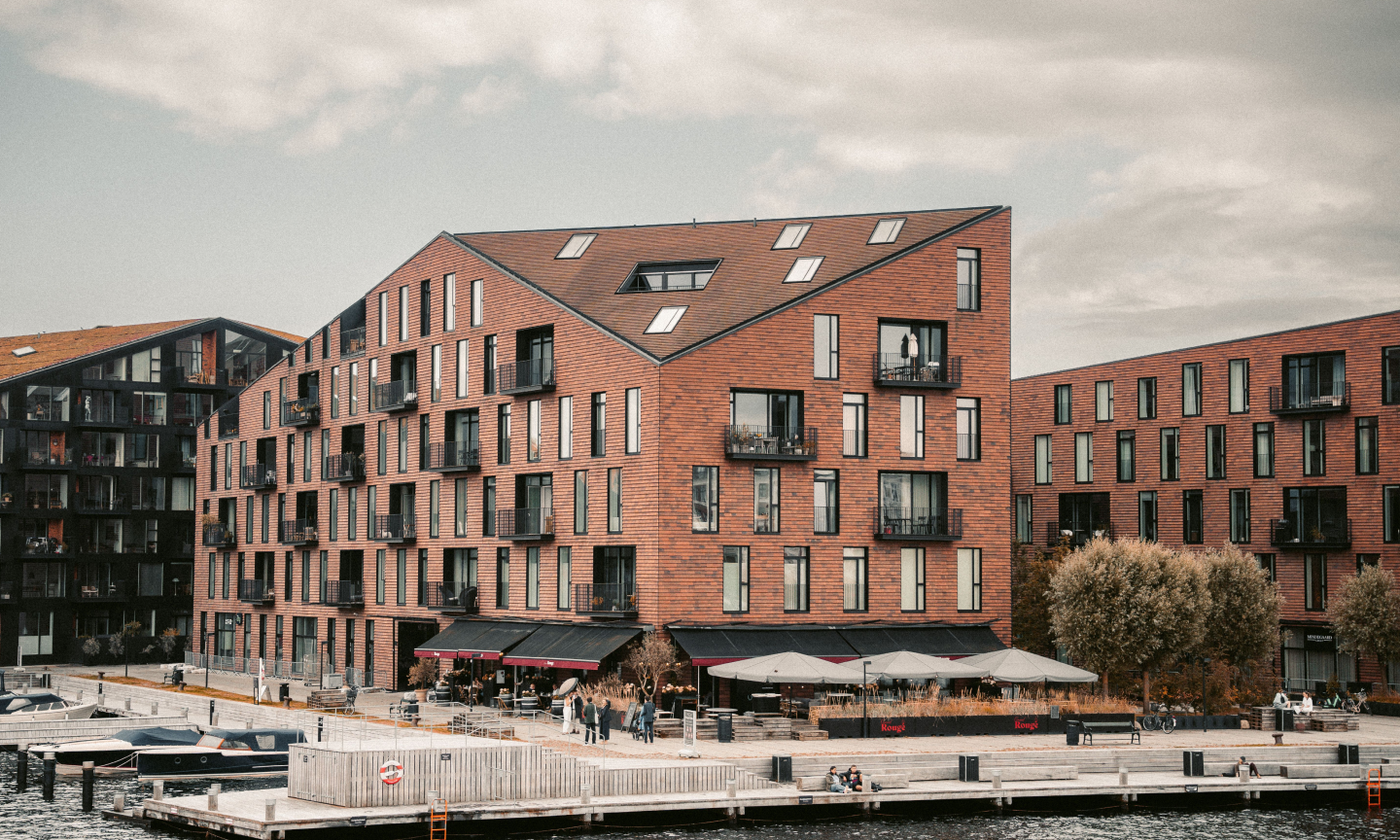 The main image for this comundo blog post about gathering data and streamlining emission data with comundo shows the Krøyers Plads residential waterfront development in the Christianshavn neighborhood of Copenhagen, Denmark, from a distance. We can see some harbour front, cafes outside the building, and a few people