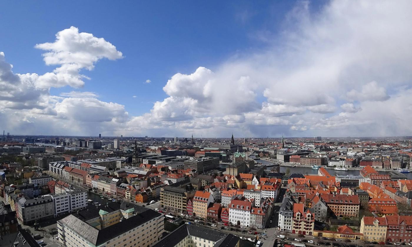 This comundo blog post has a header image that shows a bird's eye view of Copenhagen, Denmark. The city is spread out below, against a blue sky which is littered with white clouds