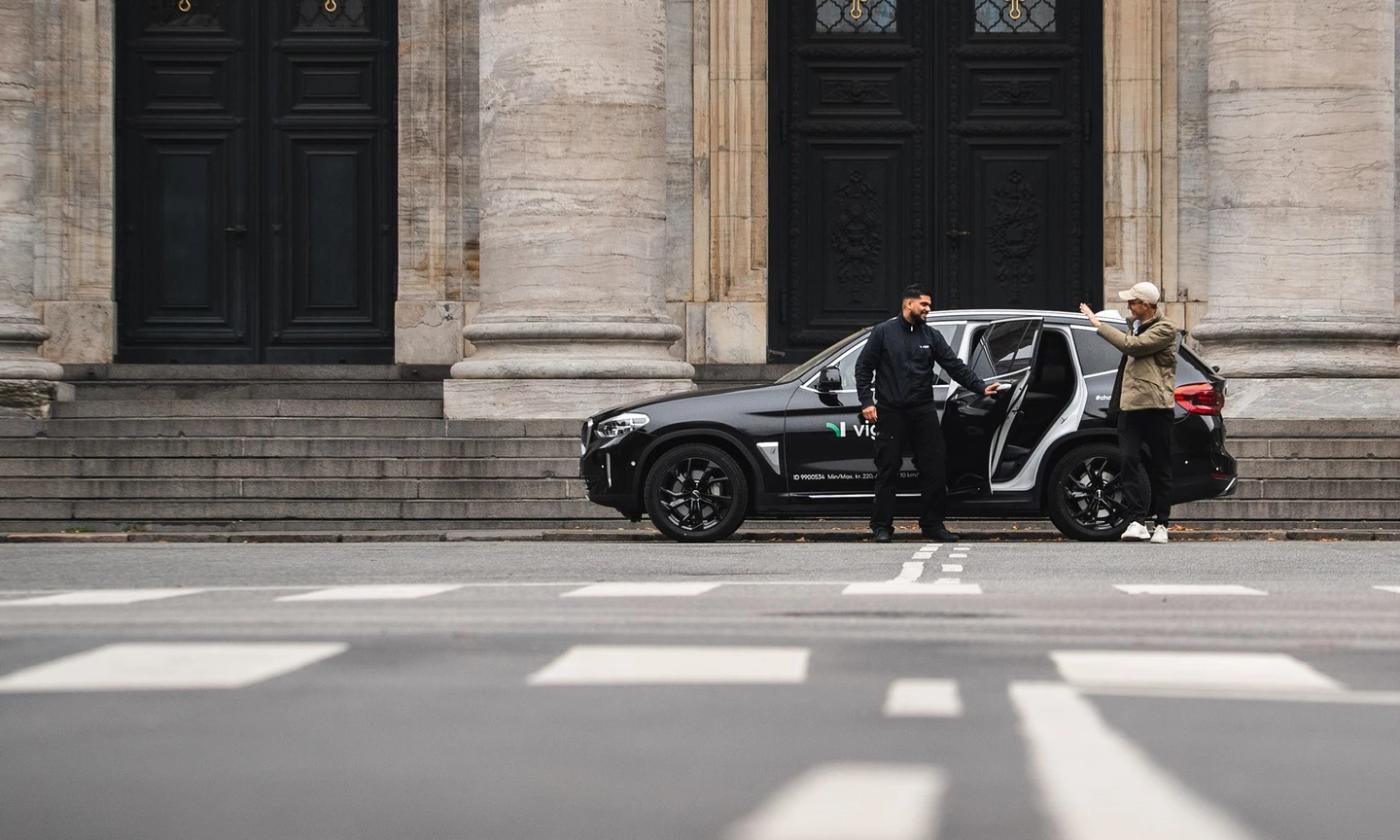 A photo of a Viggo driver opening the door of a Viggo-branded taxi parked by a large building. The passenger is out of the car and waving at the driver 