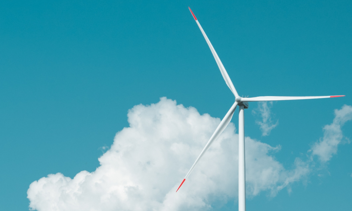 A photo of the top half of a slender wind turbine against blue sky and white fluffy clouds