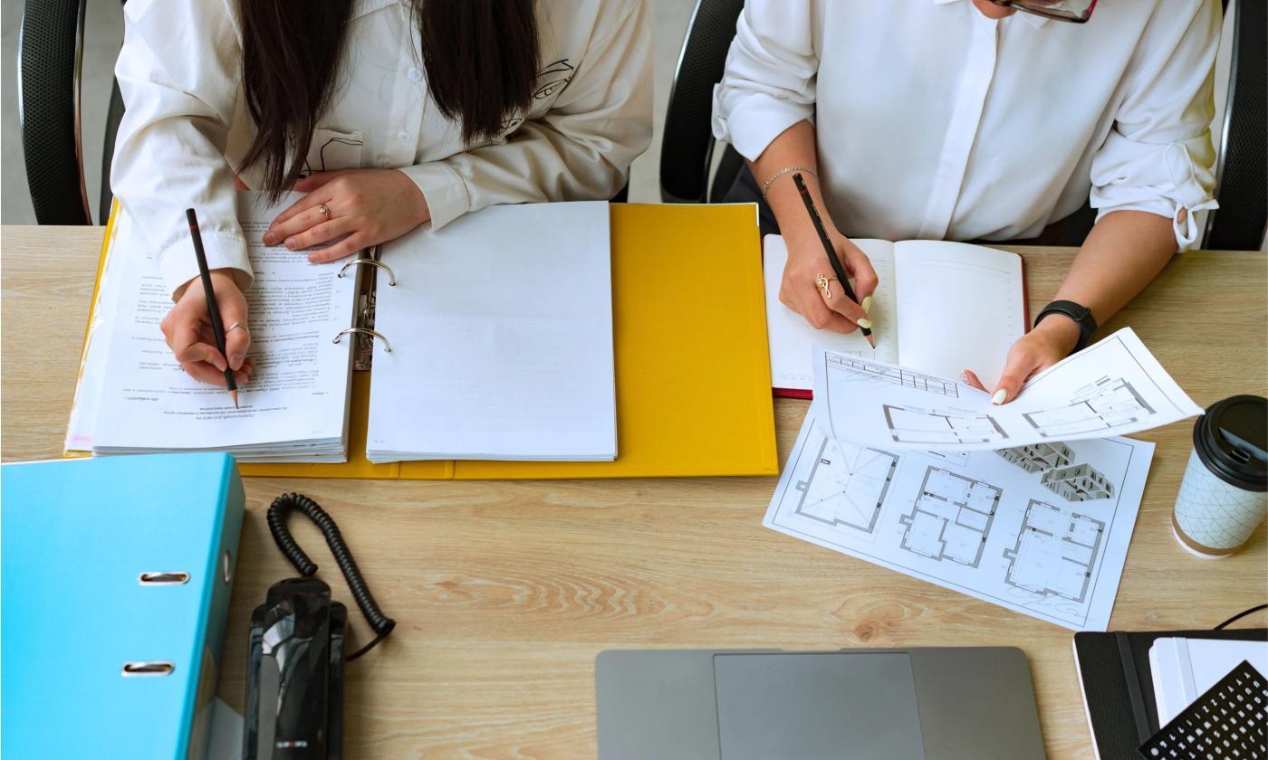 In this comundo blog post image we can see two women working from above. One has a large yellow folder open which has reams of paper in it, the other is looking at building blueprints.