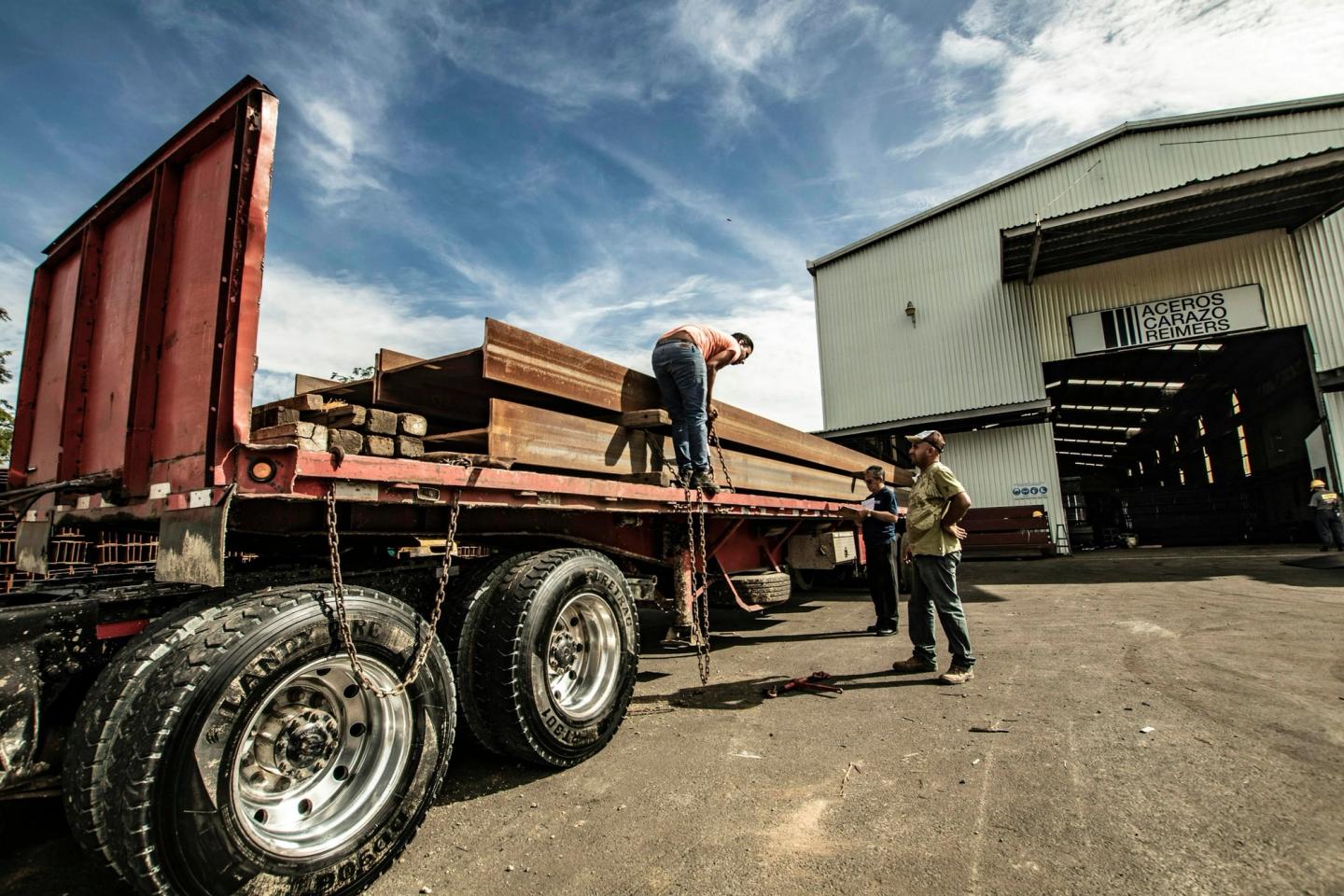 Workers unloading lumber from a flatbed trailer outside a large building.