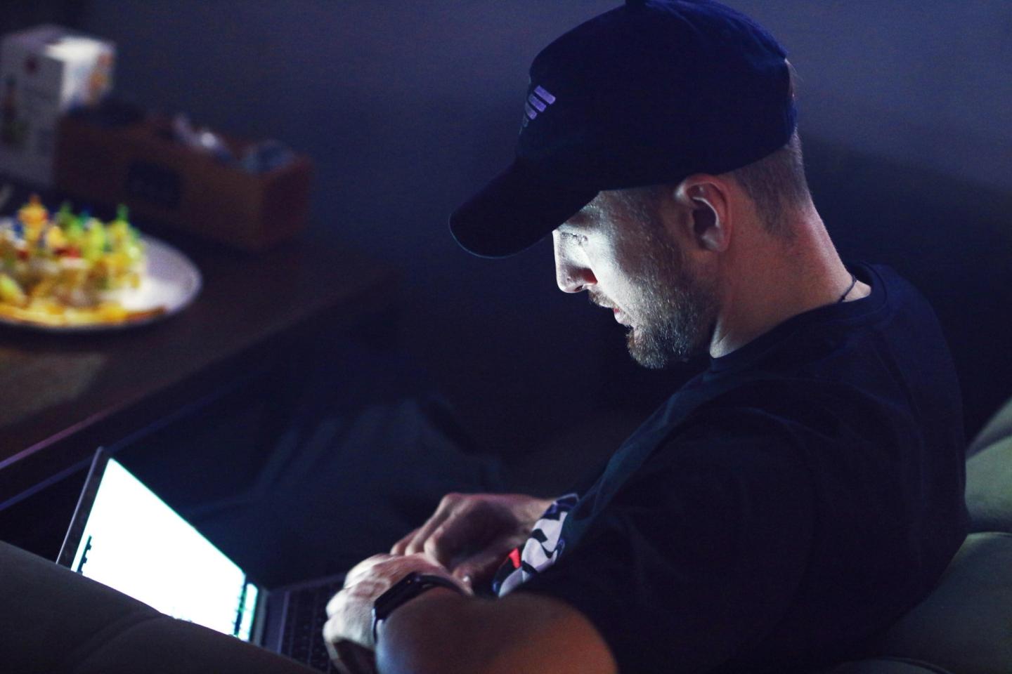 A man in a ball cap working on a laptop late in the evening.