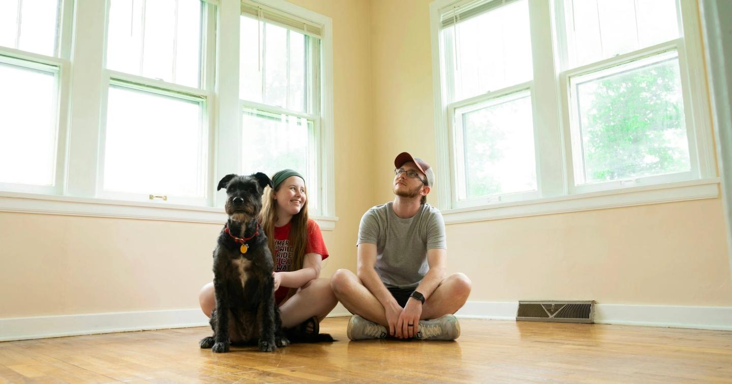 A couple and their dog sitting on the floor of an empty, sunlit room in a new home.