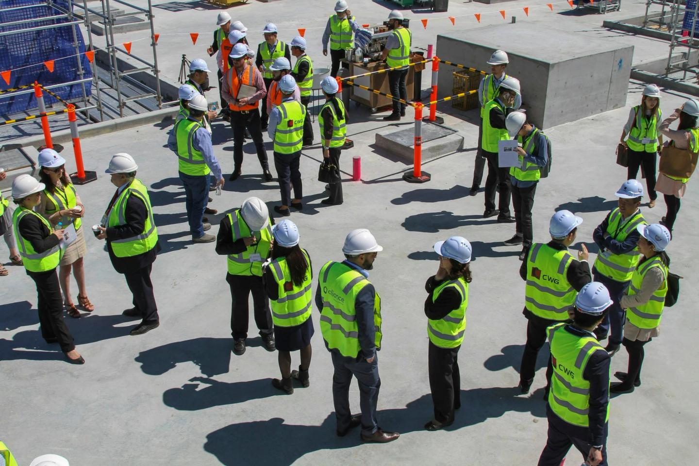 A large group of construction workers in hard hats and high-visibility vests standing together on a site for a morning huddle.