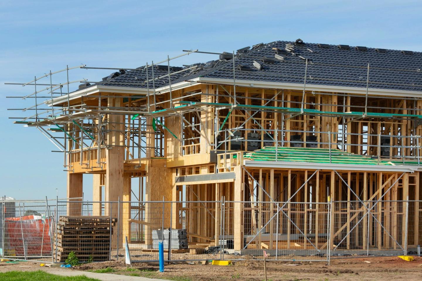 A two-storey wood-framed house going up behind a construction fence, with scaffolding and roof tiles stacked on the roof.