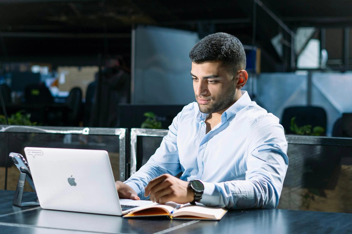 A man in a light blue shirt working on a laptop at an office desk.