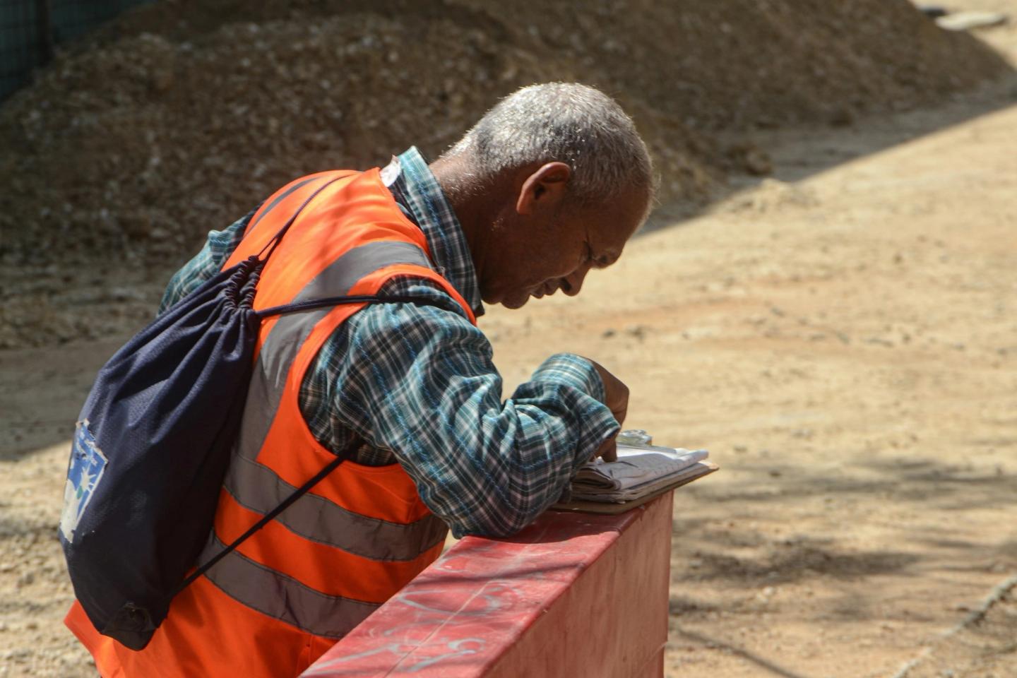 A man in a high-visibility vest bent over a notepad on a construction site, writing.