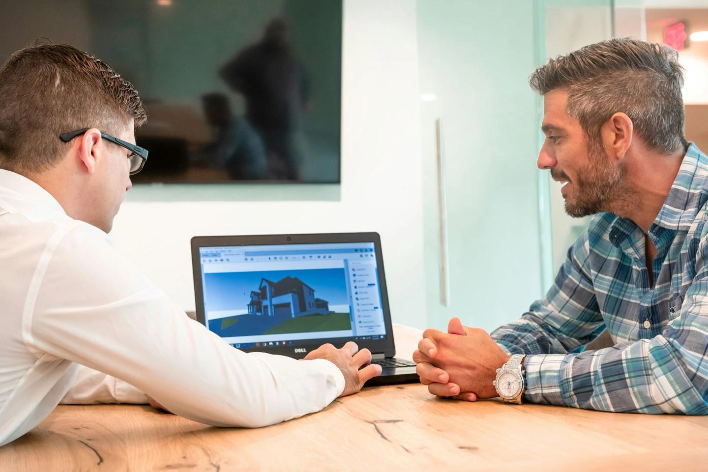 A builder and a client sitting at a table looking at a house design on a laptop.