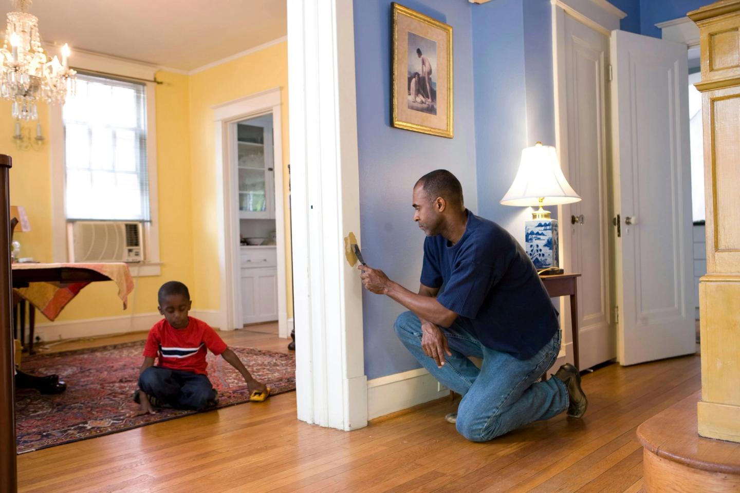A tradesman kneeling to work on a wall inside a lived-in home while a child plays on the floor.
