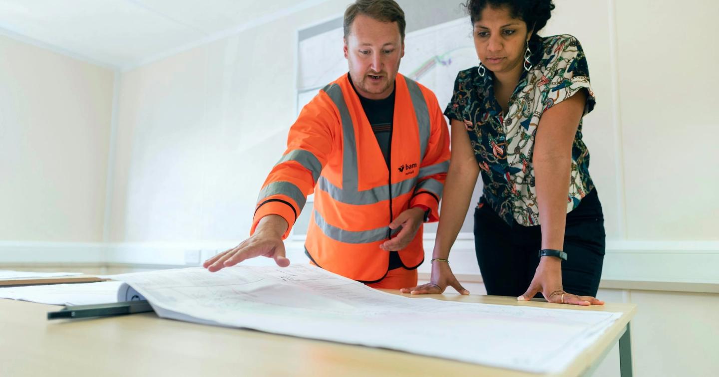 A worker in a high-visibility jacket and a colleague going over drawings on an office desk.