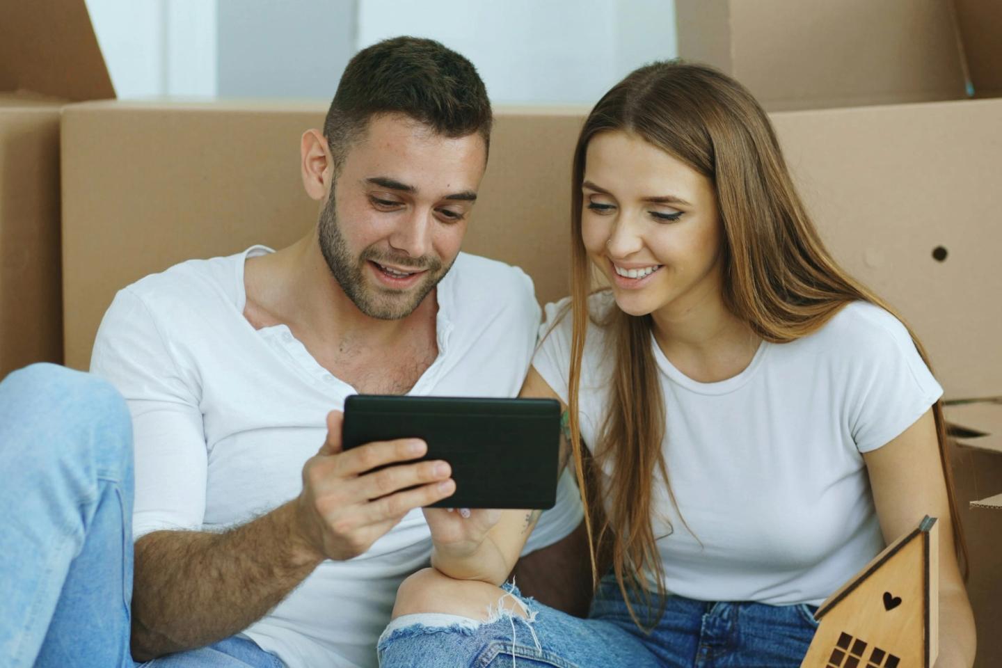 A couple sitting on the floor among moving boxes, smiling at a tablet.