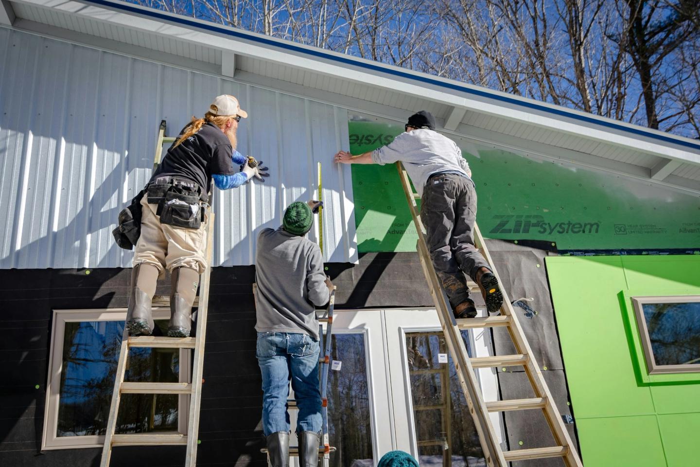 Three workers on ladders installing sheathing on the wall of a house.