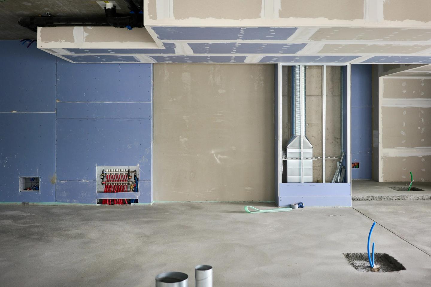 Room with drywall going up, while one wall bay still shows ductwork and floor heating pipes
