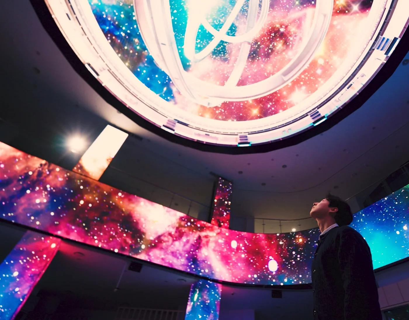 Figure gazes upward inside an immersive dome surrounded by cosmic LED displays