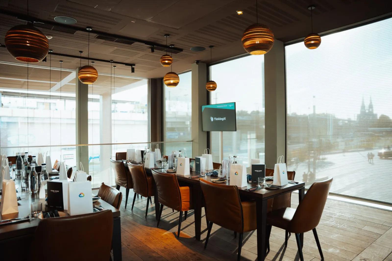 A long dinner table set with gift bags, a ThinkingAI screen and the river beyond the windows
