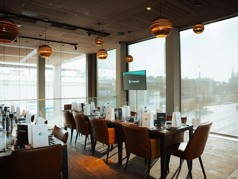 A long dinner table set with gift bags, a ThinkingAI screen and the river beyond the windows