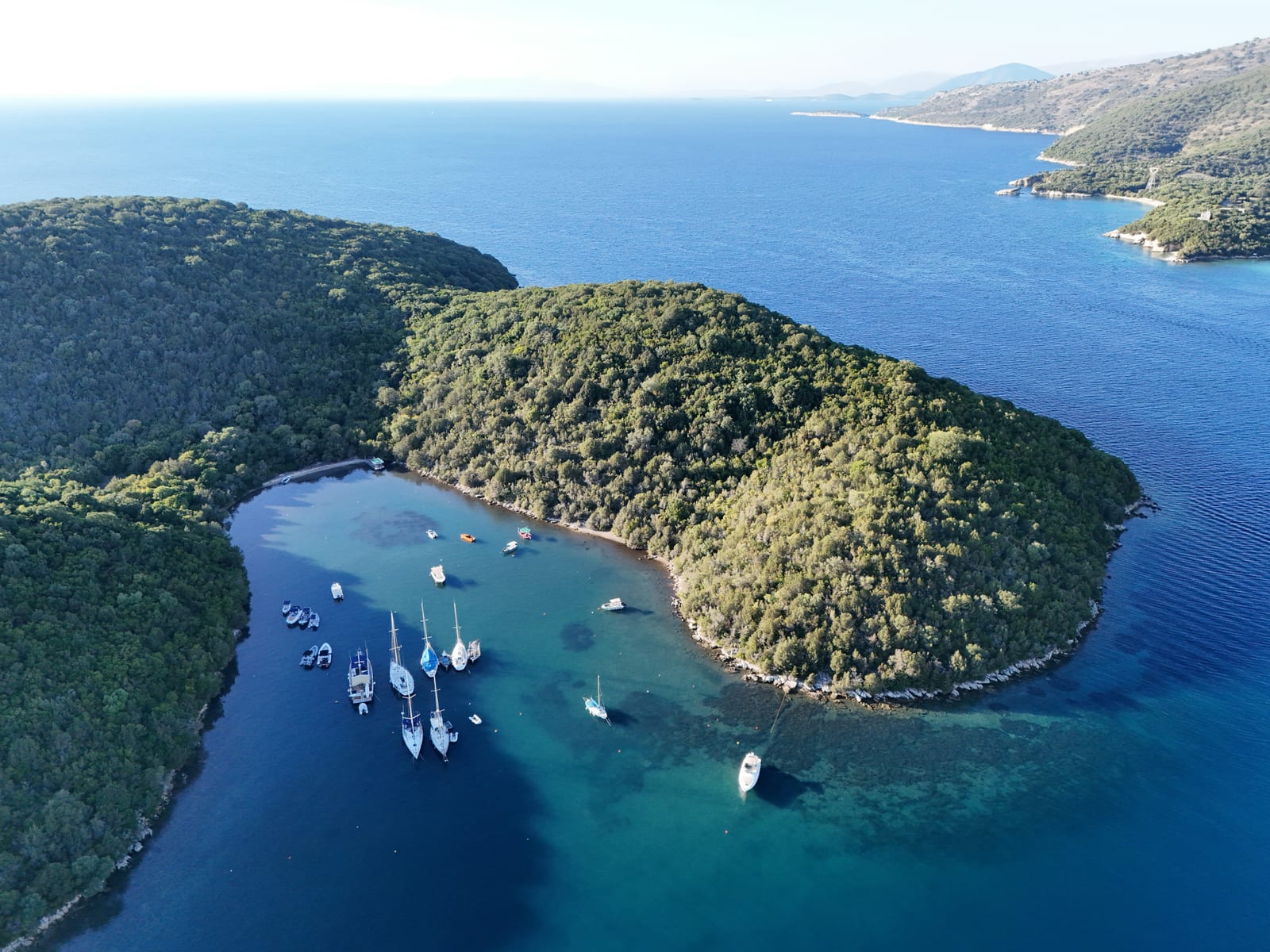 Luchtfoto van een beschutte baai met een paar zeiljachten voor anker, tussen bosrijke heuvels en helderblauw water