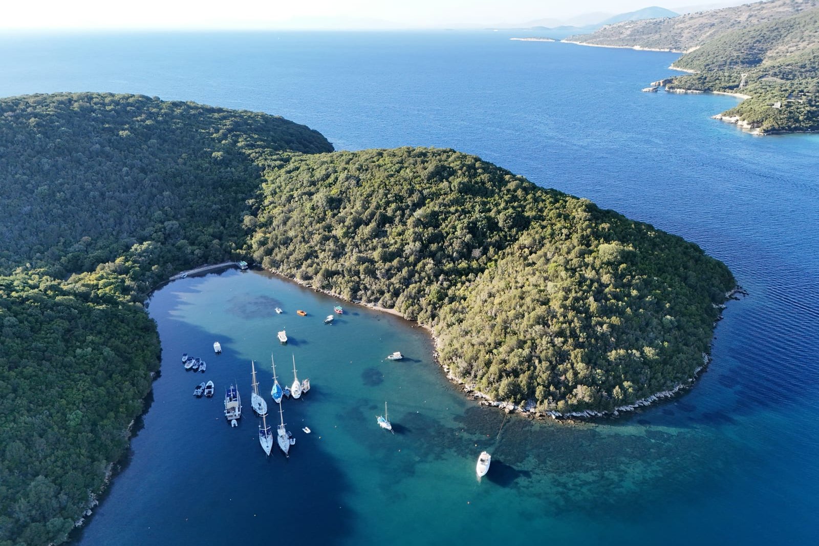 Luchtfoto van een beschutte baai met een paar zeiljachten voor anker, tussen bosrijke heuvels en helderblauw water