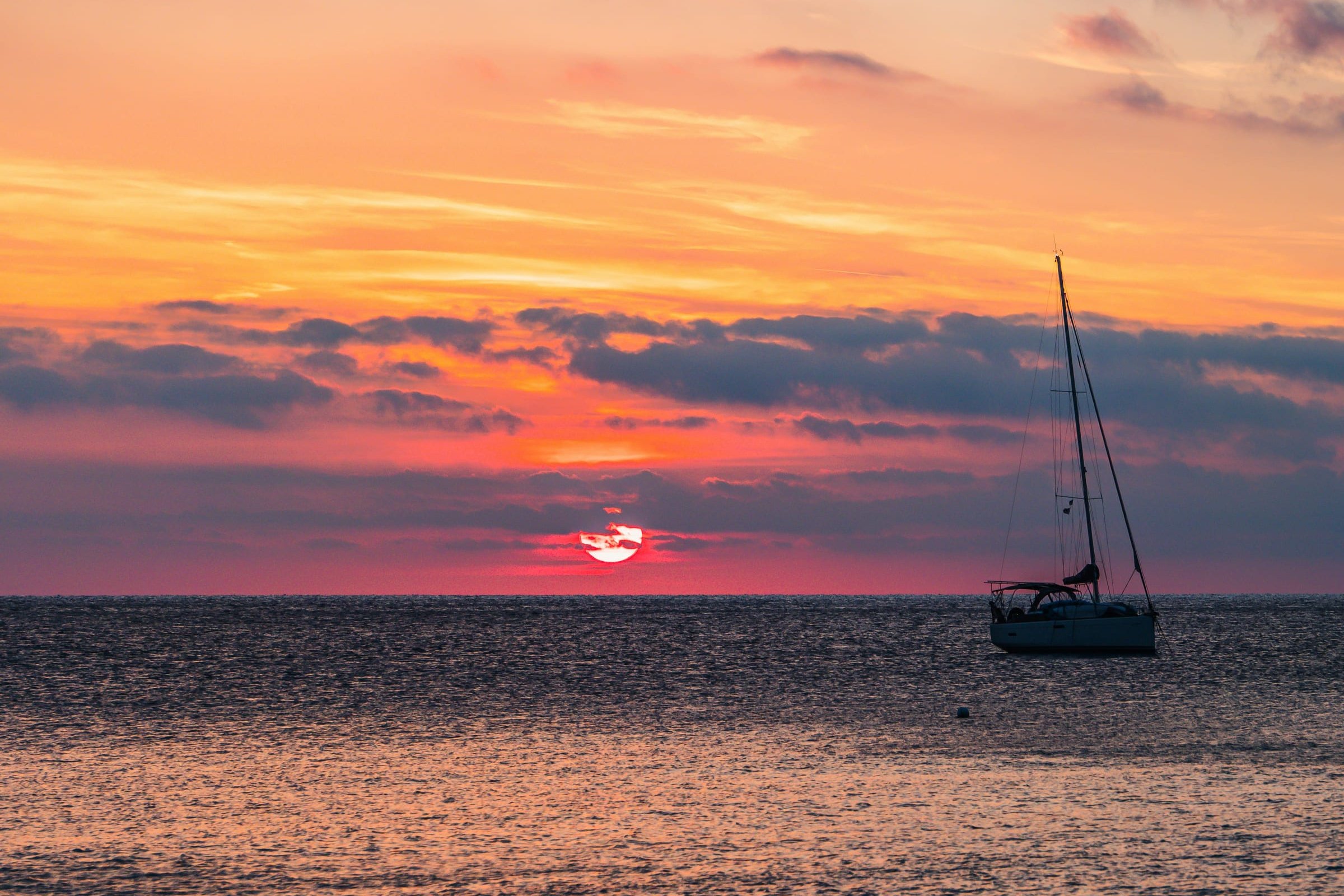 Een zeiljacht ligt voor anker op zee bij zonsondergang, onder een oranje en roze lucht