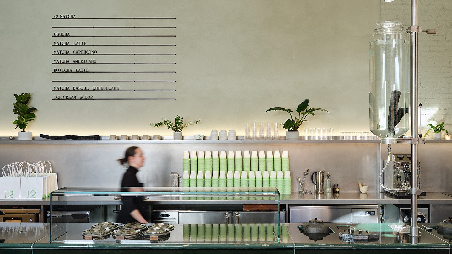 The 12Matcha café counter with a clean, industrial design. Green and white stacked cups, plants, and a minimalist menu on the wall add a fresh and organized look. A staff member moves behind the counter.