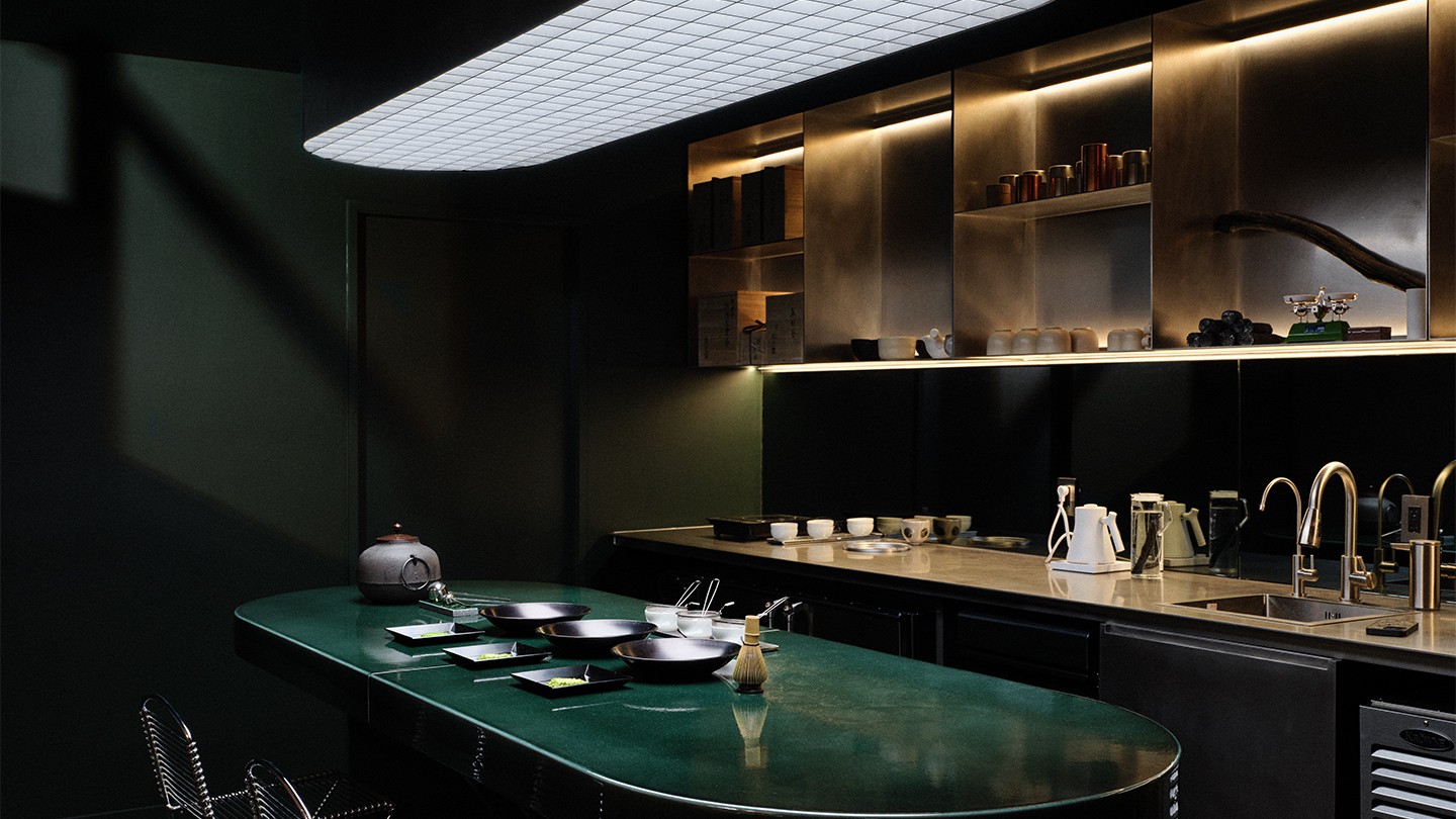 Dark, intimate tea preparation space with a green countertop and neatly arranged teaware. Soft lighting highlights the shelves filled with utensils and teacups, creating a refined and tranquil atmosphere.