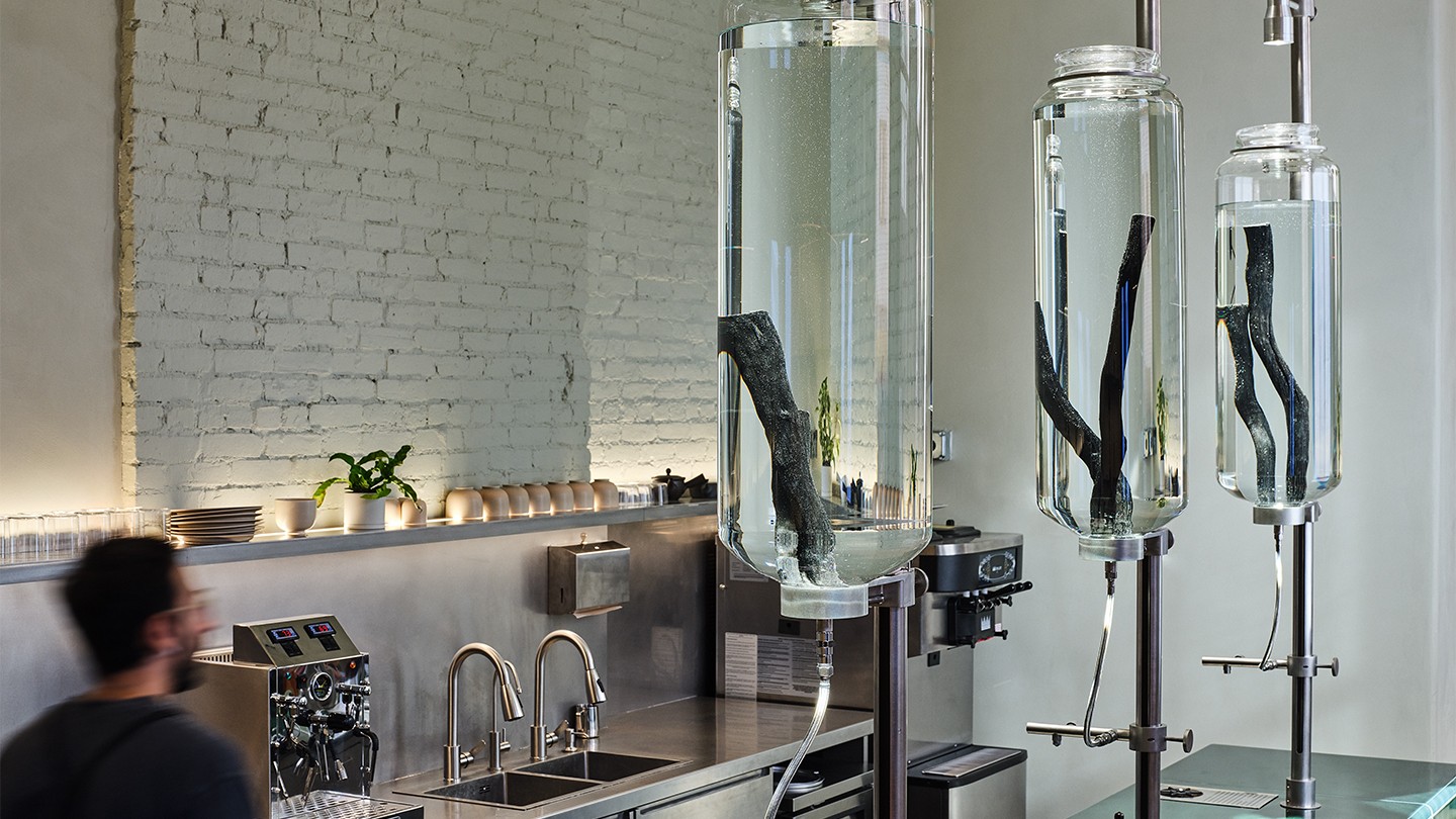 Close-up of a minimalist coffee bar featuring large cylindrical glass water drip towers with black charcoal sticks inside. Stainless steel fixtures and a clean brick wall create a sleek, modern aesthetic.
