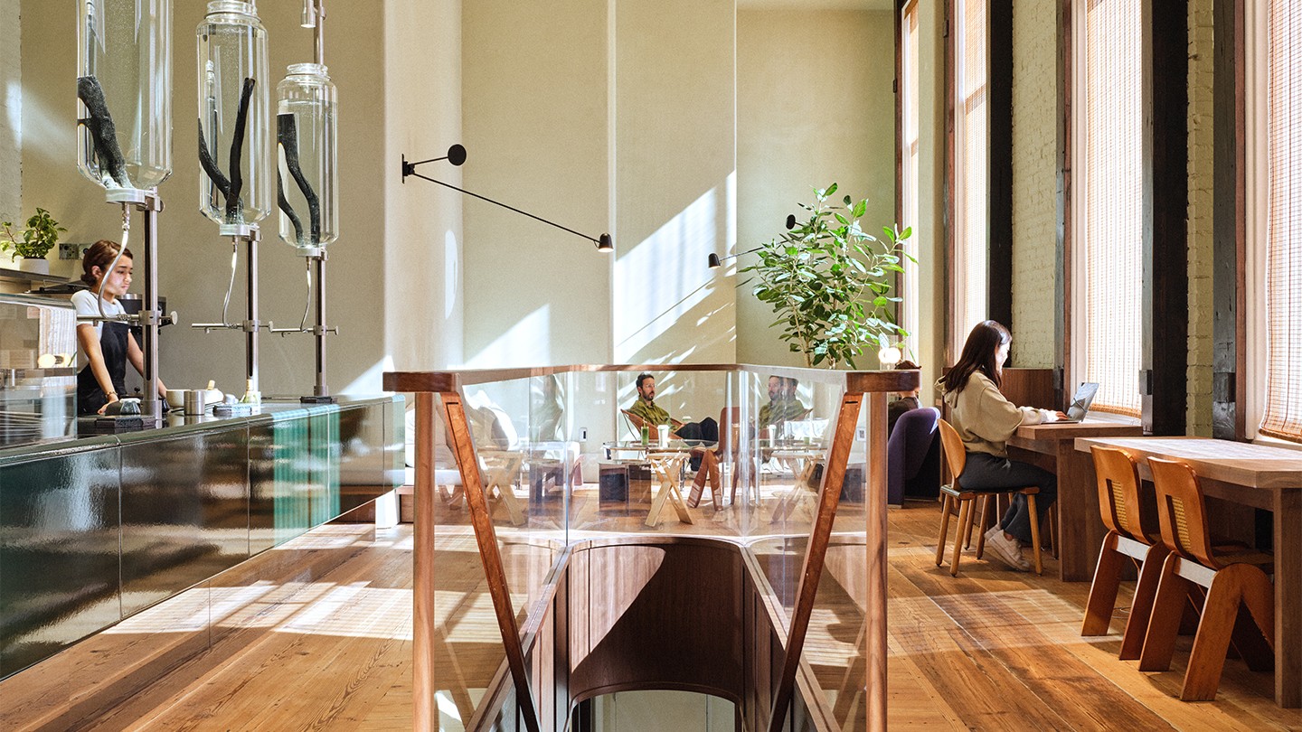 Bright and modern café interior with natural light streaming through tall windows. A barista stands behind a sleek counter with large glass brewing vessels, while customers sit at tables and workstations.