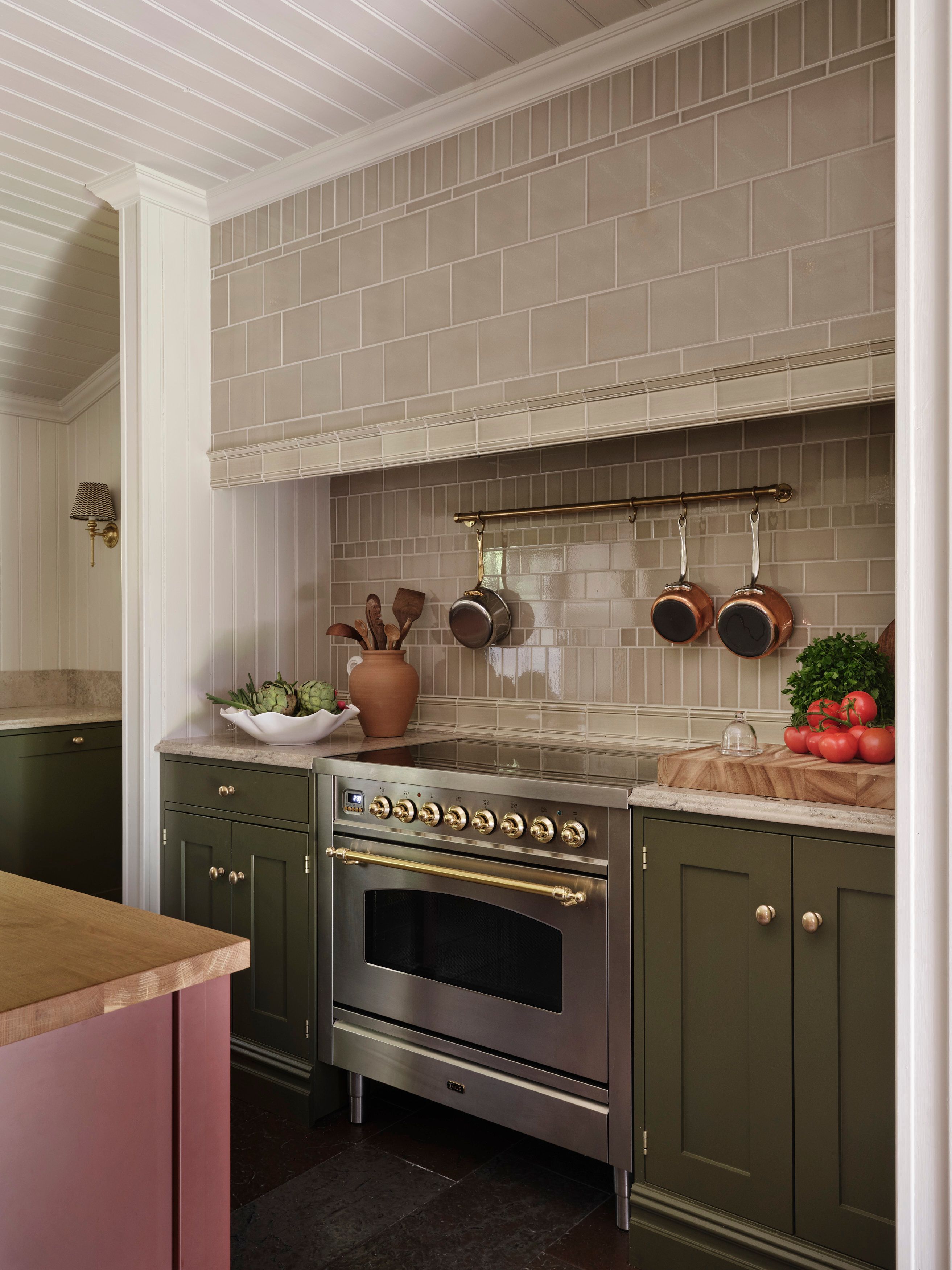 Kök med olivgröna underskåp, rostfri spis med mässingsvred och beige kakel bakom, köksö i dov rosa.